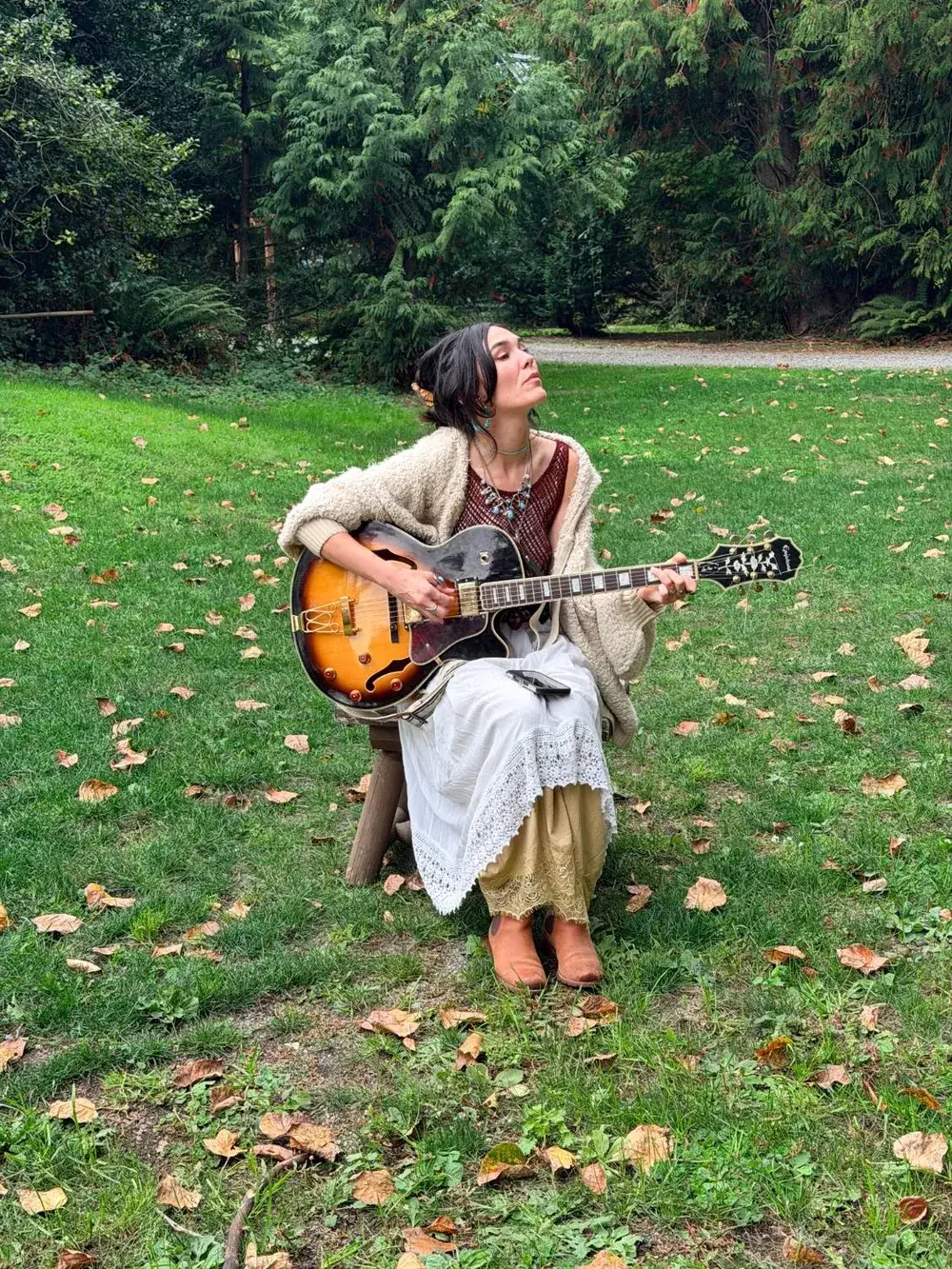 Jessie playing an archtop guitar on a stool in the grass, head tipped back, eyes closed, autumn leaves around her