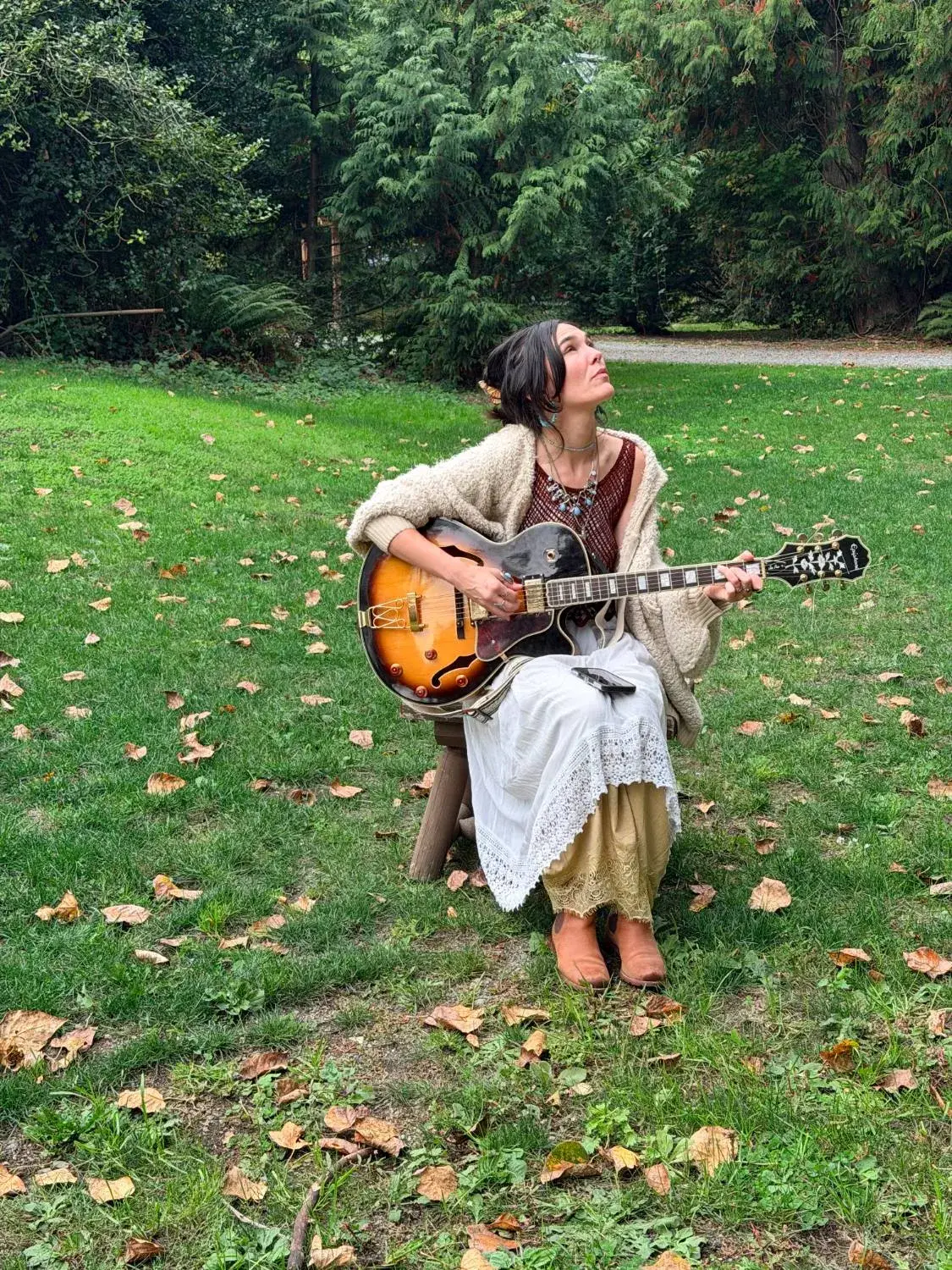 Jessie Siren sitting on a stool in a grass clearing, an archtop guitar in her lap, face turned up toward the trees